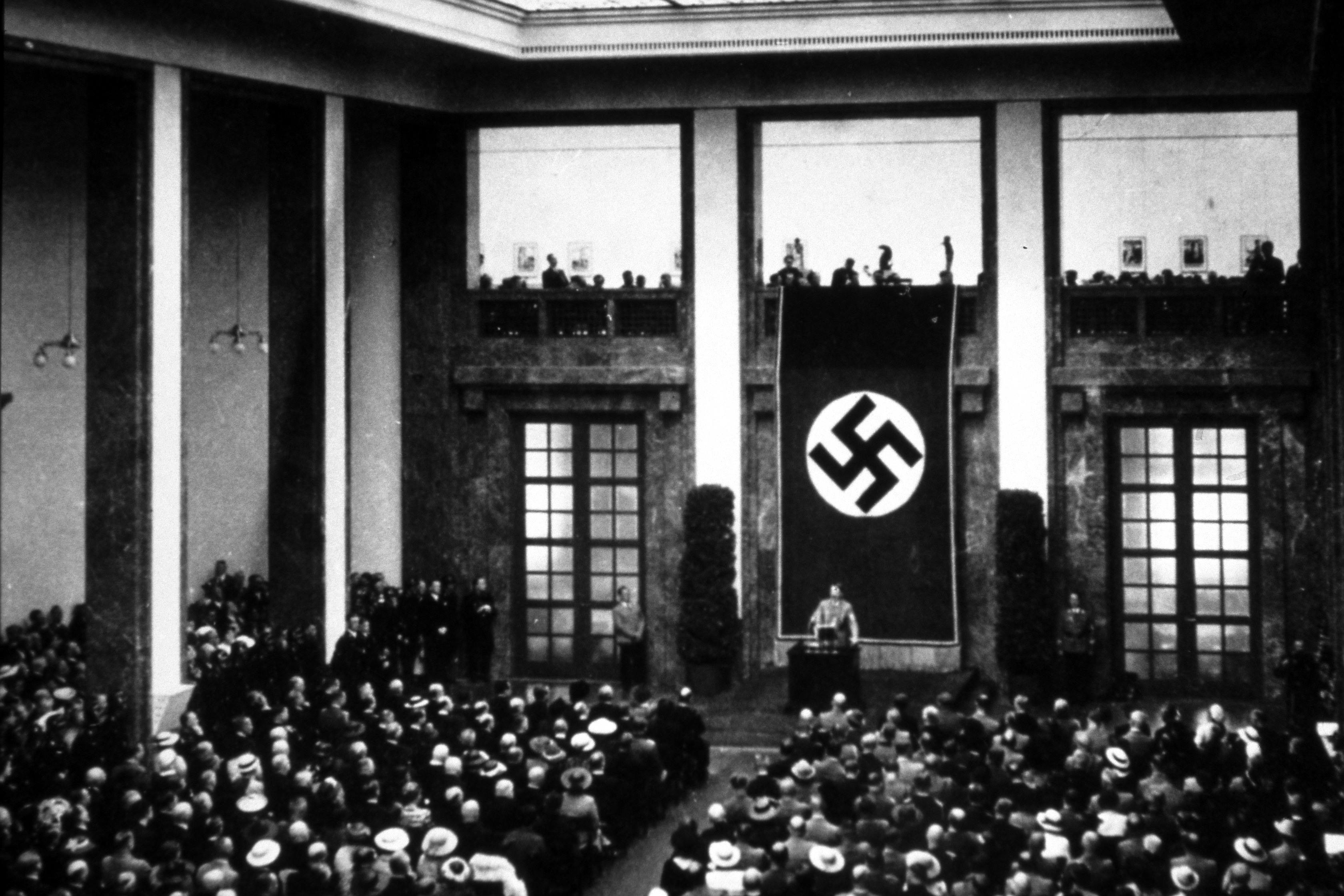 eine Menschenmasse versammelt sich vor einer Bühne in einer großen Halle. Hinter dem Redner auf der Bühne ist eine große Hakenkreuz-Flagge zu sehen. 