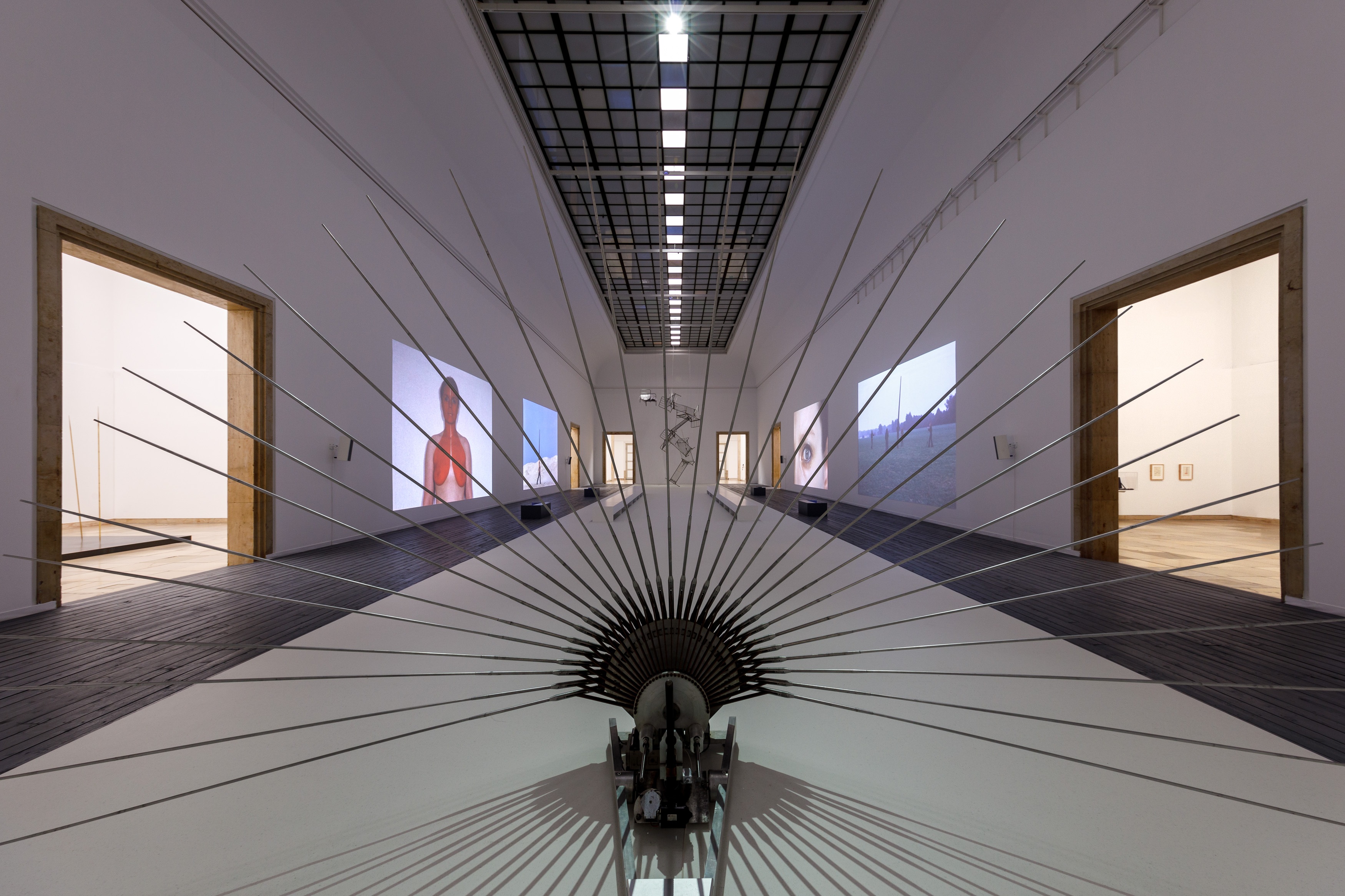 Large museum hall with film projections on the wall and a sculpture with long metal rods