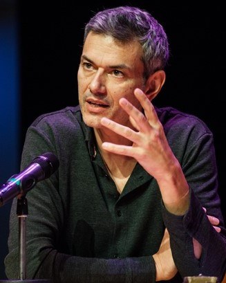 A man sits at a table and speaks into a microphone.