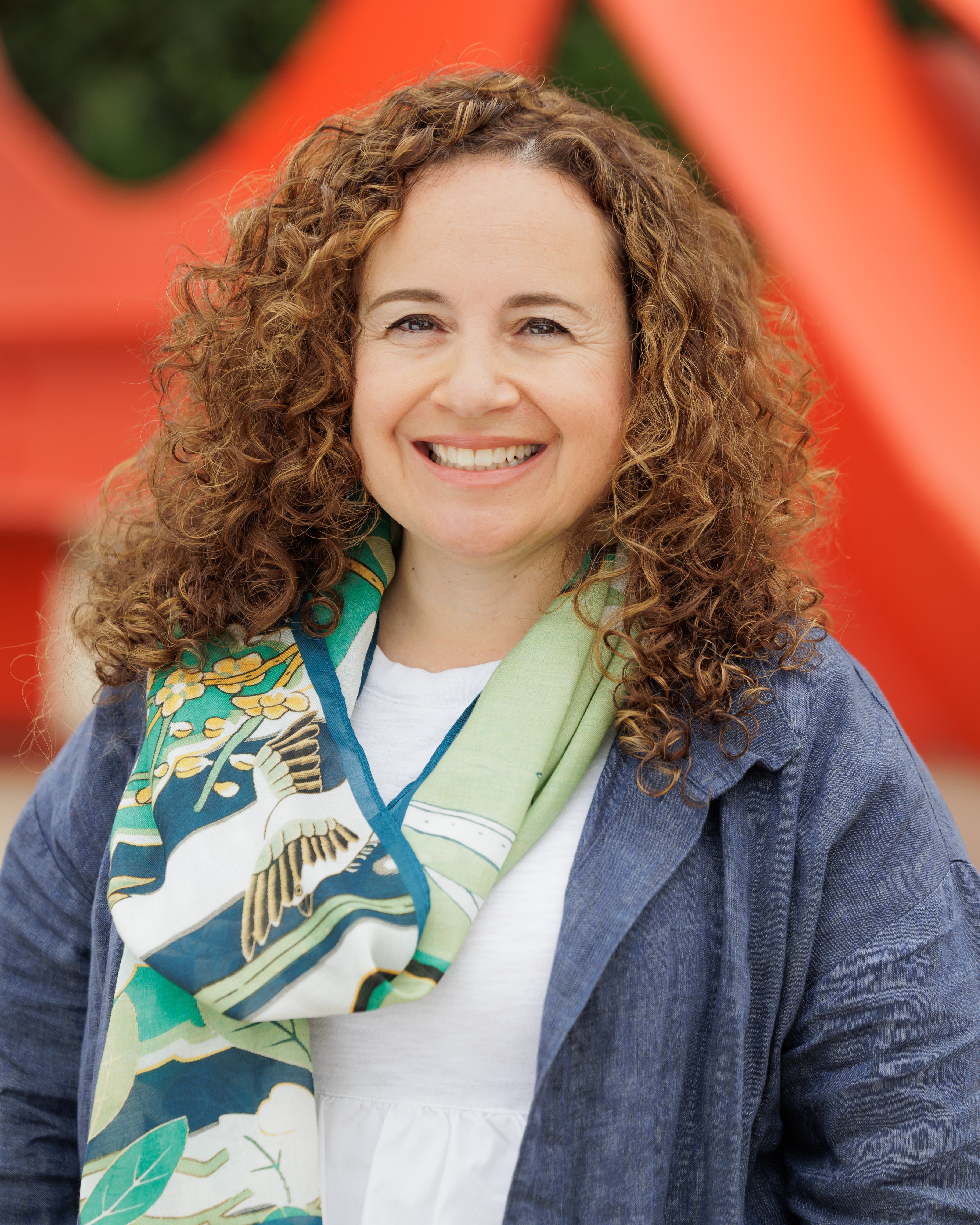 A smiling woman with light brown red curls. She is wearing a blue jacket and a green scarf.