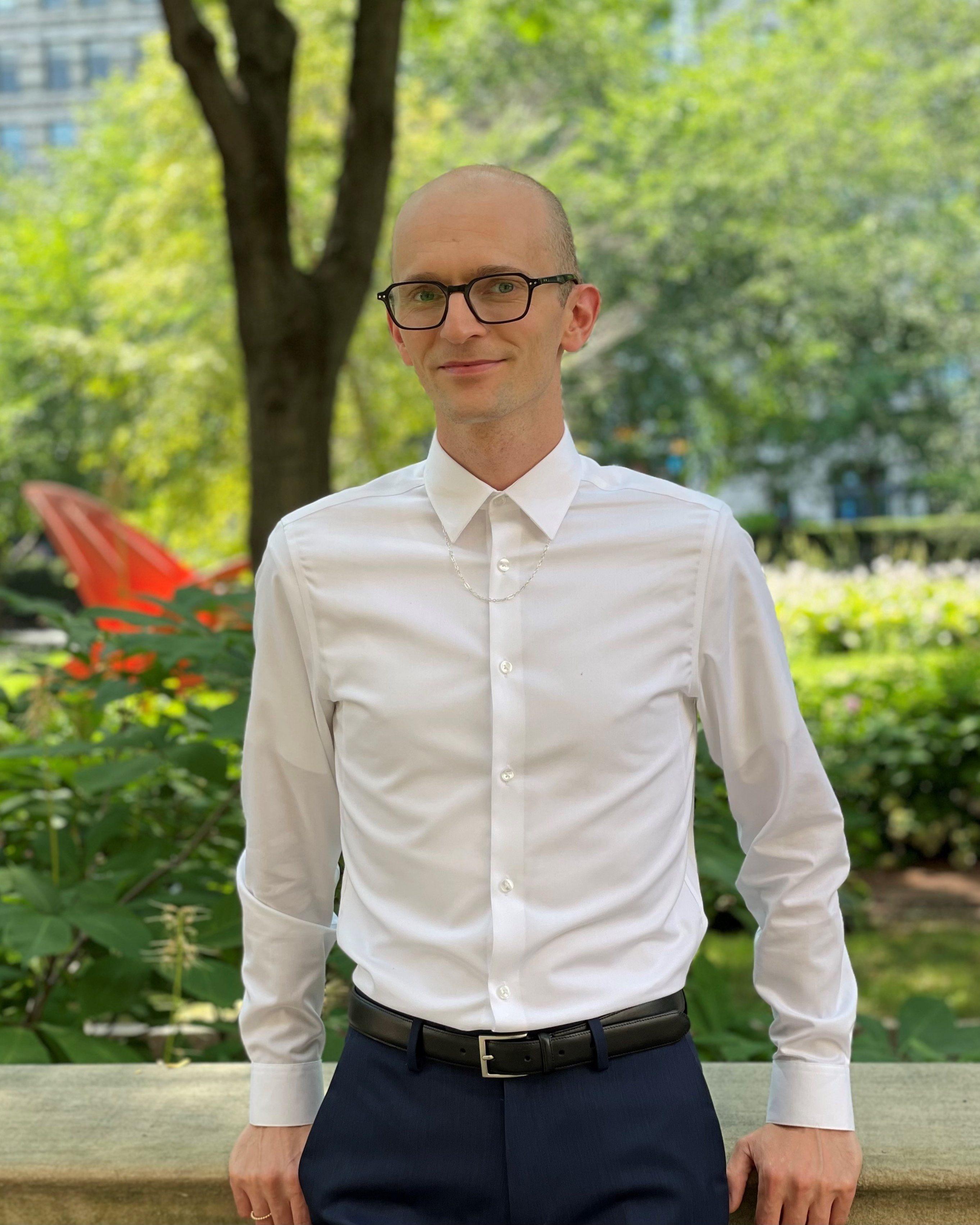 A man with a bald head and glasses is standing in front of a park. He is wearing a white shirt.