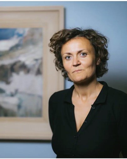 Woman with short curly hair. She is wearing a black top and standing in front of a picture.