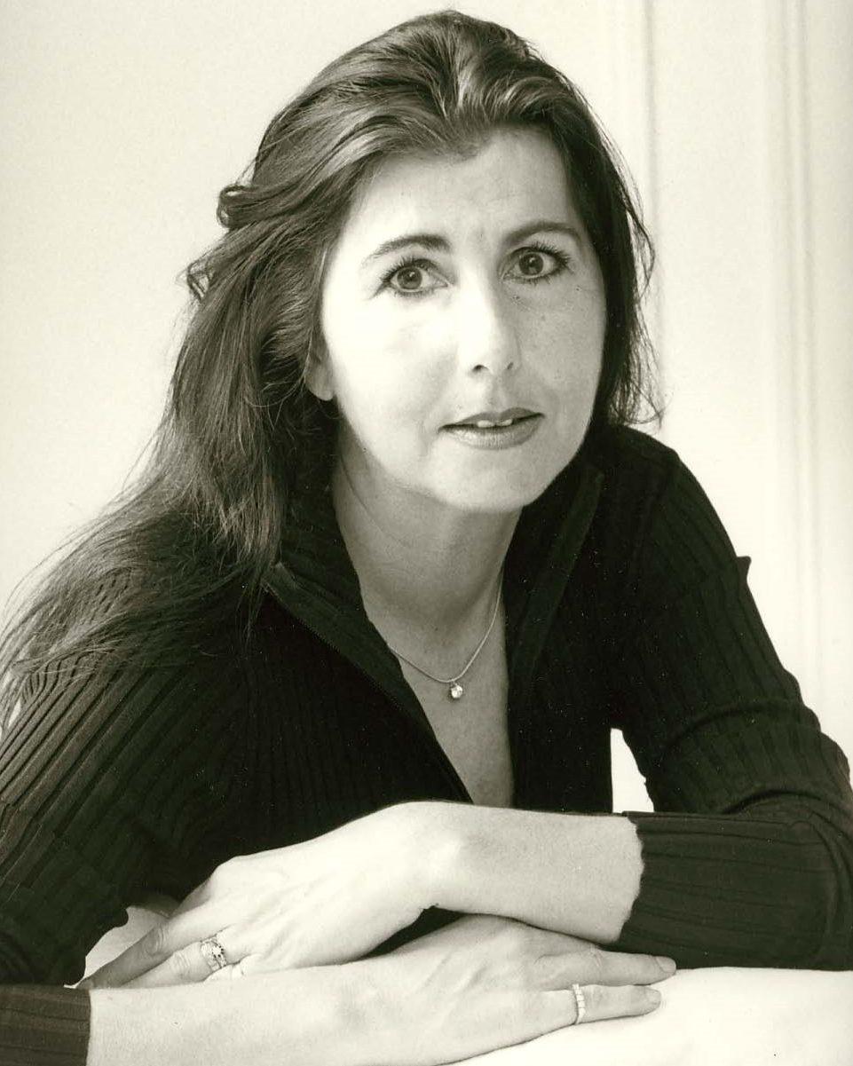 A black-and-white portrait of a woman with long, straight dark hair, a white top and standing in front of a picture.