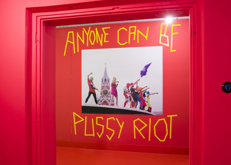 Eine rote Wand, an der ein Foto hängt. Auf dem Foto stehen mehrere Protestierende auf einem Dach. Um das Foto herum steht „Anyone can be Pussy Riot“.