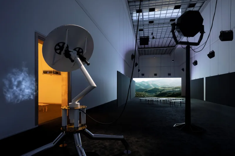 Eine Raumansicht mit einem großen Screen, Lautsprechern an der Decke und einer sateligtenartige Skulptur.