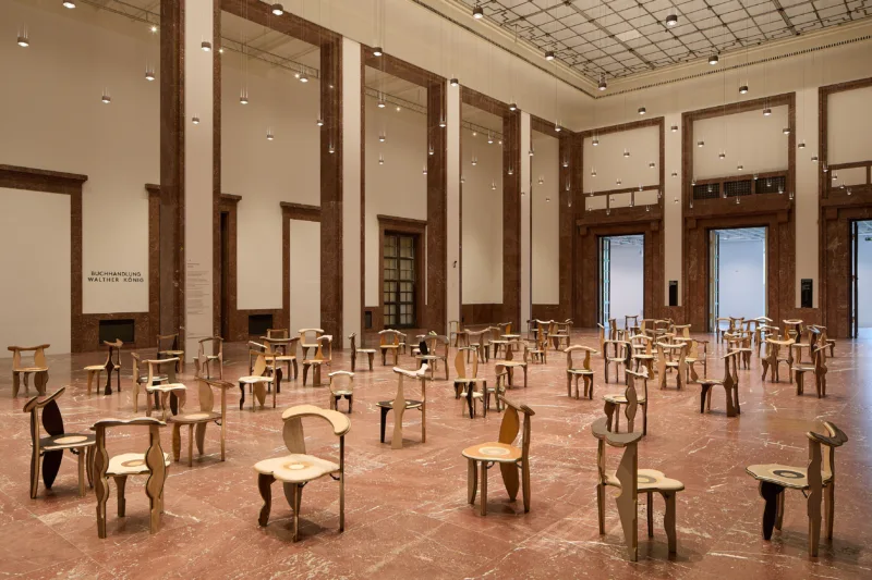 In einer großen Halle stehen zahlreiche individuelle Stühle aus Holz verteilt auf dem rot-braunen Marmorboden.