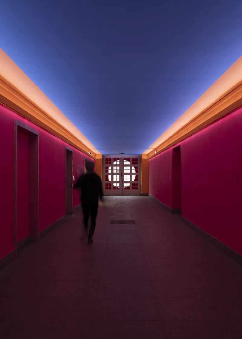 Orange erleuchteter langer Raum mit pinken WÄnden und blauer Decke. Am Ende des Raums ist eine Tür mit einem großen kreisförmigen Fenster.