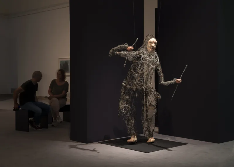 Ausstellungsansicht mit einer menschenähnlichen Skulptur aus Draht mit einer weißen Maske als Kopf.