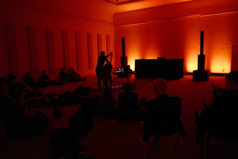 Ein dunkler Raum mit rotem Licht. Eine Person spielt ein Instrument. Eine Gruppe von Zuhörern.