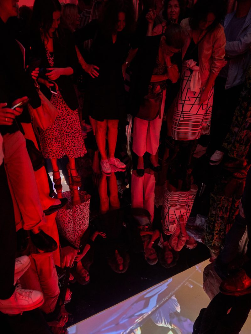 Menschen stehen in einem rot beleuchteten Raum auf einem verspiegelten Boden.