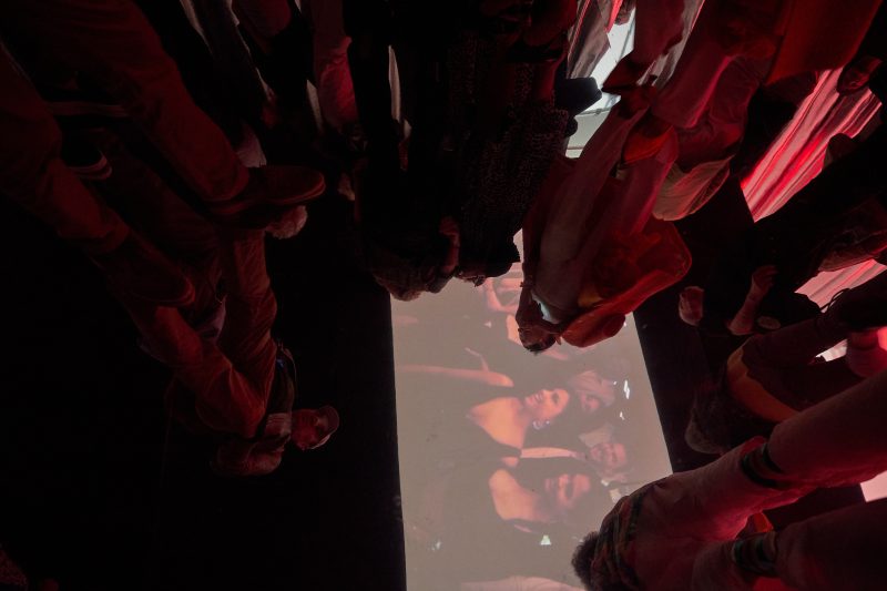 Stehende Gruppe von Menschen, von unten fotografiert. Videoprojektion an der Decke.