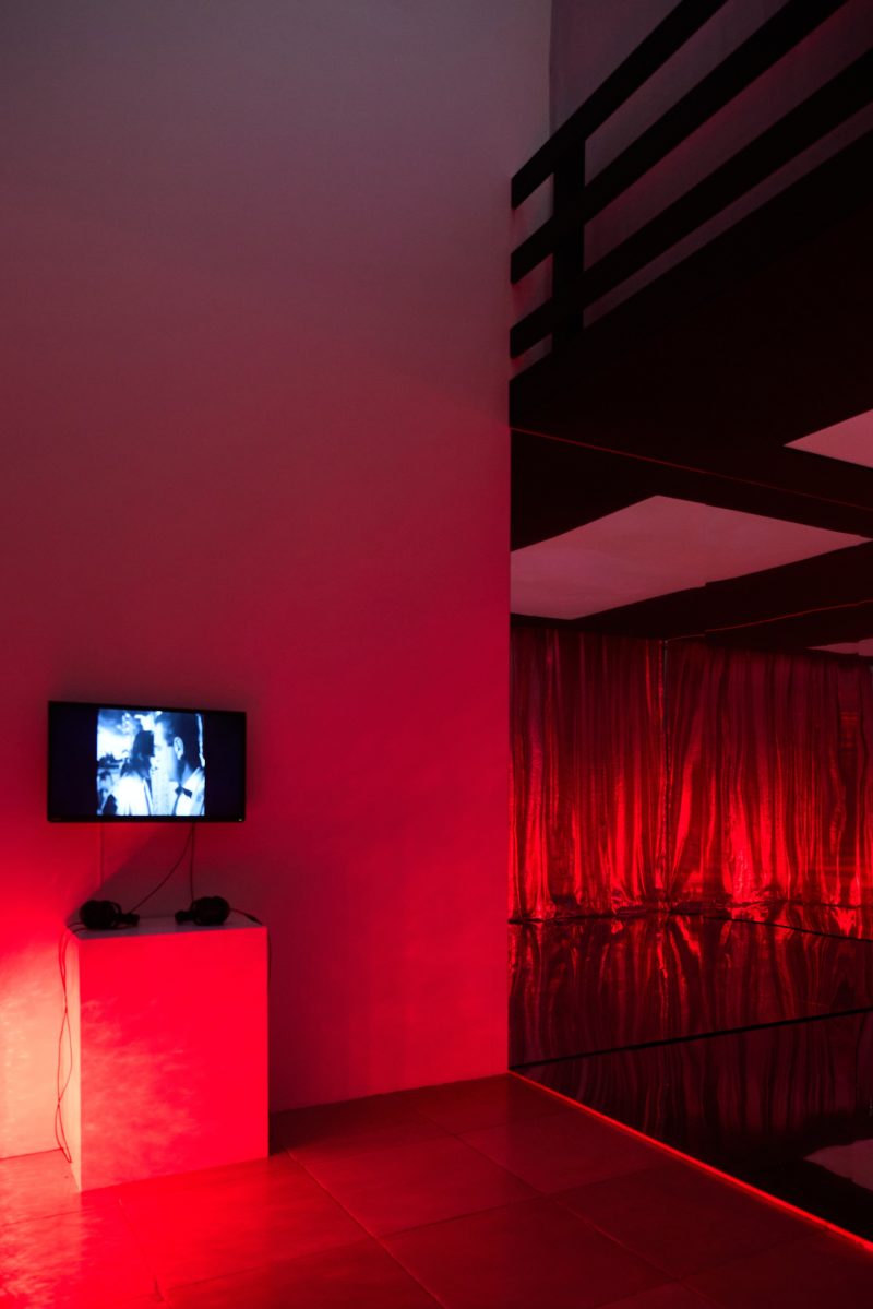 Rot beleuchteter Raum mit einem kleinen Monitor an der Wand, der ein Video abspielt.