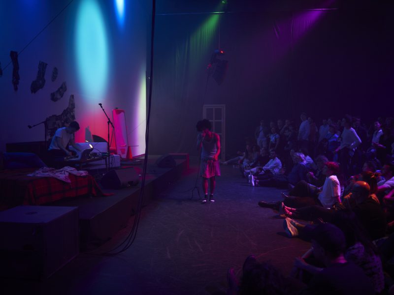 Ein dunkler Raum mit bunten Akzenten. Eine Person singt auf der Bühne vor Zuschauenden.