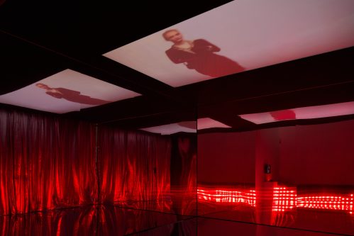 Rot beleuchteter Raum mit verspiegeltem Boden, verspiegelten Wänden und Vorhängen. Videoprojektionen an der Decke.