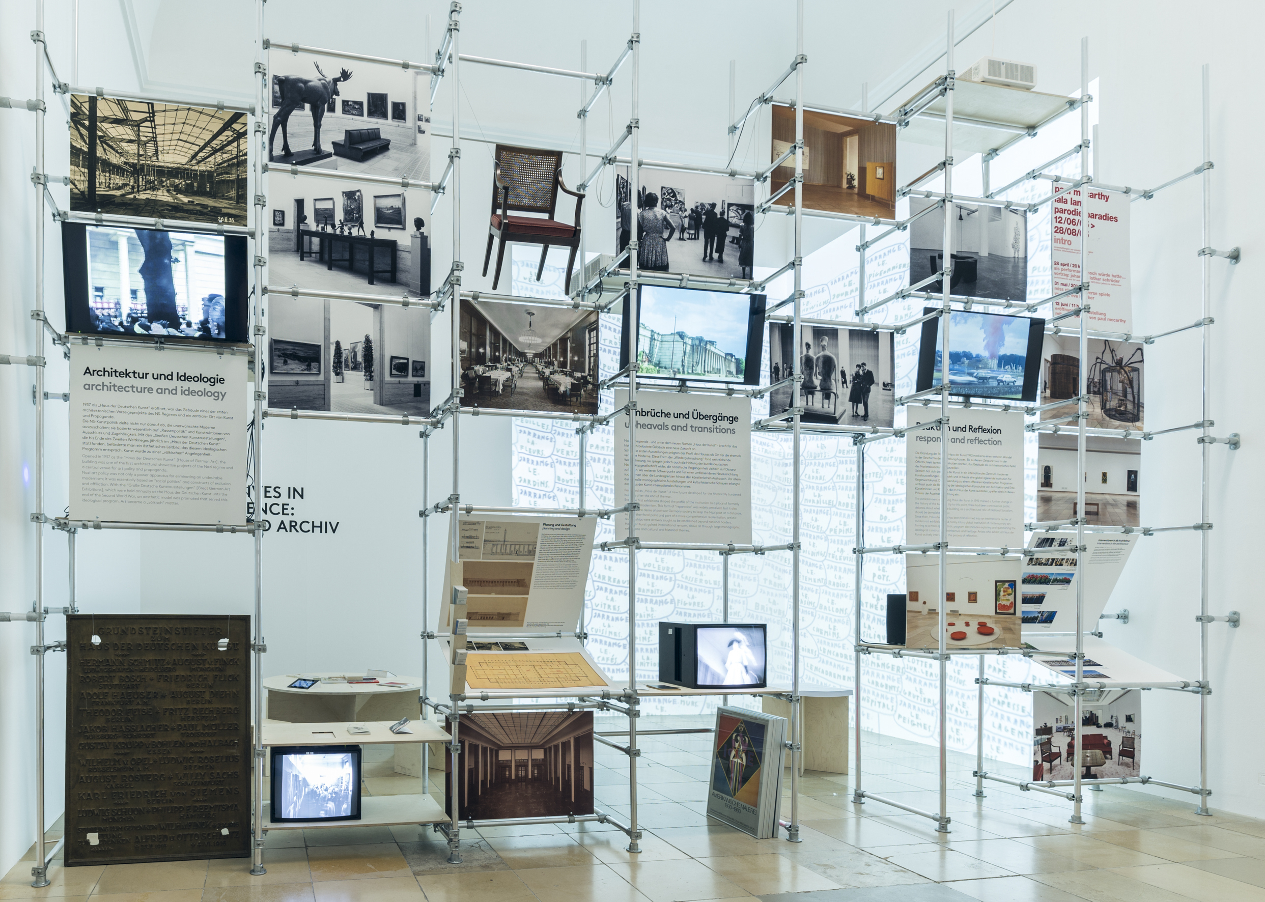 A scaffolding made of metal tubes containing screens, photographs, panels and a chair. 